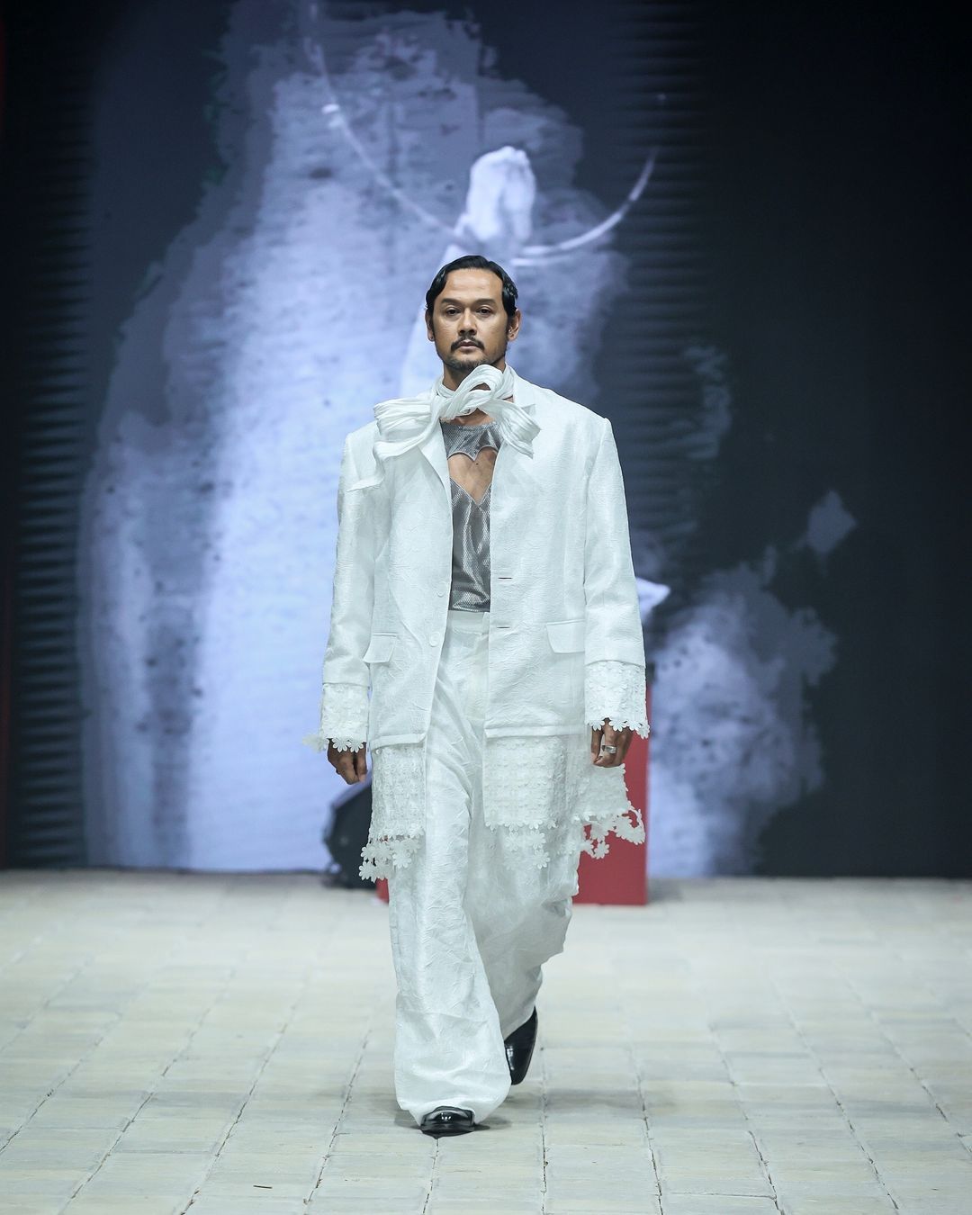 danjyo hiyoji hadirkan esensi kabaret yang semarak di pimfw 2024, 1. Koleksi ‘Monsieur’ dibuka dengan deretan look yang menampilkan setelan blazer yang pas dan celana panjang yang bervolume. Aksesori scarf dengan konstruksi kaku yang seolah tertiup angin memberi dimensi ekstra yang unik di sejumlah look., 2. Koleksi ini menangkap sensasi dari pertunjukan kabaret dengan menata ulang siluet busana pria klasik dengan volume yang berlebihan dan lapisan yang didekonstruksi., 3. Potongan asimetris dan kain berlapis menciptakan kontras visual yang mencolok. Elemen-elemen yang ceria seperti lengan yang besar dan motif yang dramatis menambahkan sentuhan unik yang memberi kejutan dalam koleksi ini., 4. Dwi Sasono jadi salah satu muse yang berjalan di runway, memamerkan koleksi setelan jas dengan sentuhan elemen floral dan celana serba putih yang dipadukan dengan inner warna silver dan scarf yang dirancang dengan konstruksi kaku., 5. Adinia Wirasti muncul dengan penampilan yang ikonik seperti di film The Greatest Showman. Mengenakan setelan jas yang mencolok dengan siluet yang dramatis, mewujudkan daya tarik kabaret yang serba glamor.