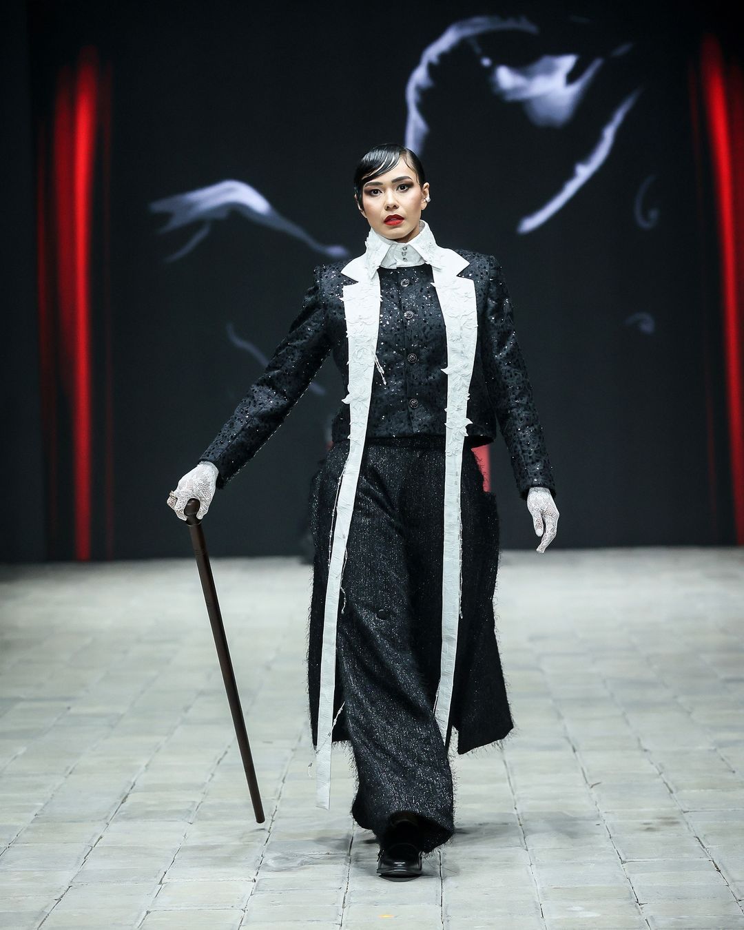 danjyo hiyoji hadirkan esensi kabaret yang semarak di pimfw 2024, 1. Koleksi ‘Monsieur’ dibuka dengan deretan look yang menampilkan setelan blazer yang pas dan celana panjang yang bervolume. Aksesori scarf dengan konstruksi kaku yang seolah tertiup angin memberi dimensi ekstra yang unik di sejumlah look., 2. Koleksi ini menangkap sensasi dari pertunjukan kabaret dengan menata ulang siluet busana pria klasik dengan volume yang berlebihan dan lapisan yang didekonstruksi., 3. Potongan asimetris dan kain berlapis menciptakan kontras visual yang mencolok. Elemen-elemen yang ceria seperti lengan yang besar dan motif yang dramatis menambahkan sentuhan unik yang memberi kejutan dalam koleksi ini., 4. Dwi Sasono jadi salah satu muse yang berjalan di runway, memamerkan koleksi setelan jas dengan sentuhan elemen floral dan celana serba putih yang dipadukan dengan inner warna silver dan scarf yang dirancang dengan konstruksi kaku., 5. Adinia Wirasti muncul dengan penampilan yang ikonik seperti di film The Greatest Showman. Mengenakan setelan jas yang mencolok dengan siluet yang dramatis, mewujudkan daya tarik kabaret yang serba glamor.